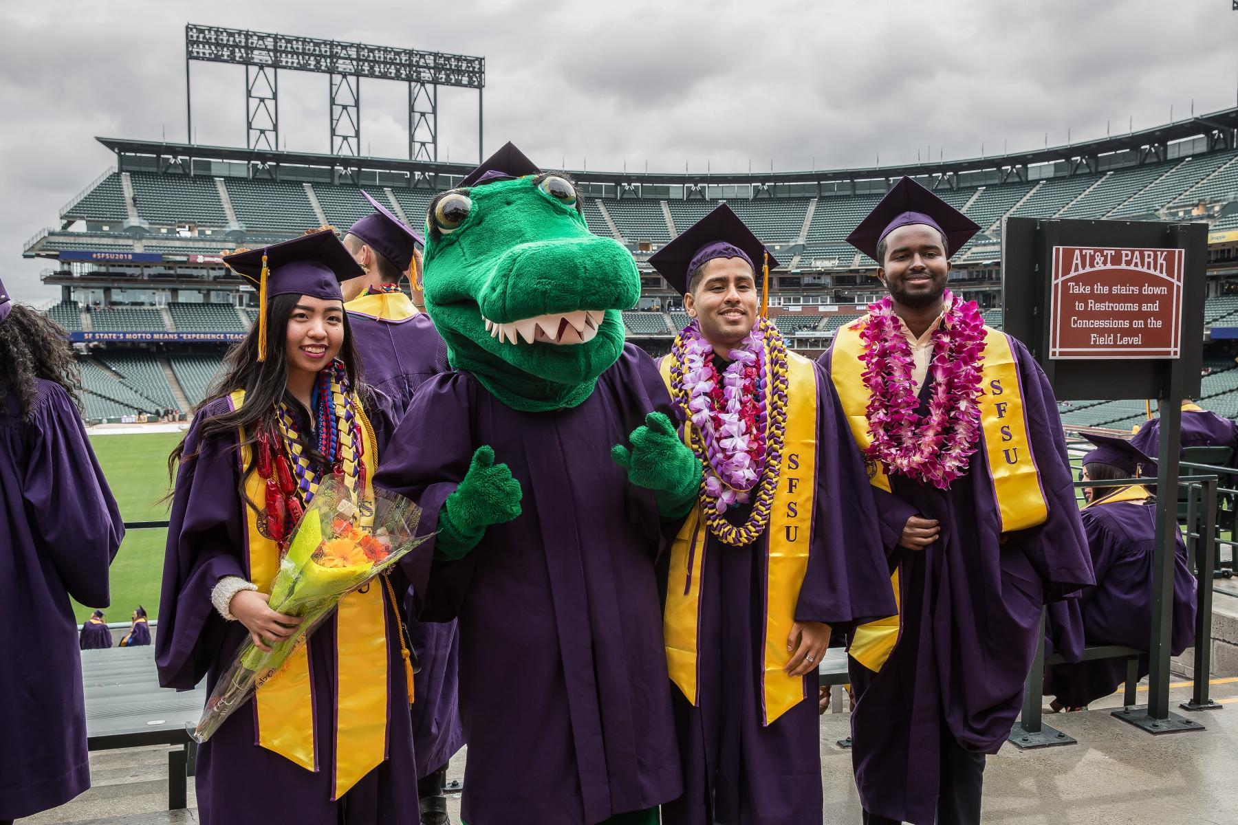 Three students dressed in graduation regalia pose with the Gator mascot at AT&T park.